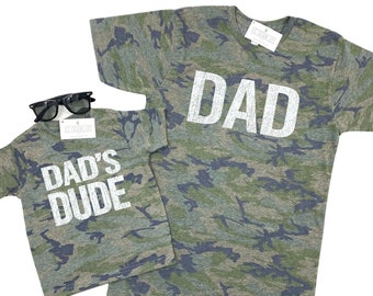 May include: Two camouflage t-shirts with white text. The larger shirt says "DAD" and the smaller shirt says "DAD'S DUDE".