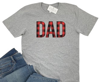 May include: A gray t-shirt with the word "DAD" in a red and black buffalo plaid pattern. The shirt is displayed with a pair of blue jeans. A tag is visible in the upper right corner.