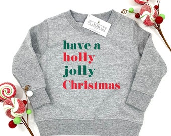 May include: A gray sweatshirt with the text "have a holly jolly Christmas" in green and red. The sweatshirt has a crew neck and long sleeves. A tag is attached to the collar. Decorative candy canes and berries are in the background.