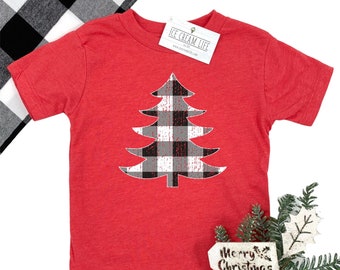 May include: A red short-sleeved t-shirt featuring a black and white buffalo plaid Christmas tree design. The shirt has a tag that says "ICE CREAM LIFE". A black and white checkered fabric is visible in the background. A sign that says "Merry Christmas" is in the foreground.