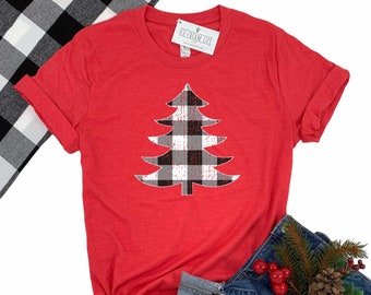 May include: A red t-shirt featuring a Christmas tree design in a black, white, and gray buffalo plaid pattern. The shirt has rolled-up sleeves and is paired with blue jeans and a black and white checkered cloth.