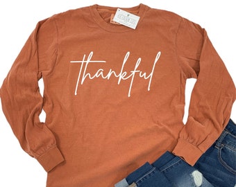 May include: A long-sleeved, rust-colored t-shirt with the word "thankful" written in white script on the front. The shirt is folded and displayed on a white background.