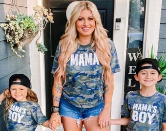 Könnte beinhalten: Eine Frau und zwei Kinder tragen passende T-Shirts mit Camouflage-Print. Auf dem Shirt der Frau steht "MAMA" in weißen Buchstaben. Auf den Shirts der Kinder steht "MAMA'S BOY". Die Frau trägt Jeansshorts. Die Kinder tragen Jeans und Baseballkappen.