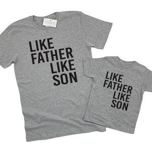 May include: Two grey t-shirts with black text that reads "Like Father Like Son". The larger shirt has the text stacked vertically, while the smaller shirt has the text stacked horizontally.