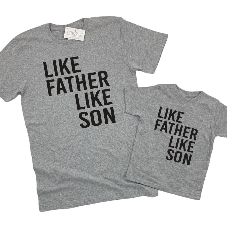 May include: Two grey t-shirts with black text that reads "Like Father Like Son". The larger shirt has the text stacked vertically, while the smaller shirt has the text stacked horizontally.