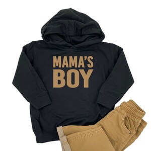 May include: Black hooded sweatshirt with the text "MAMA'S BOY" in brown letters. The sweatshirt is paired with brown pants.