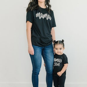 Pode incluir: Uma m&atilde;e e sua filha vestindo camisetas pretas combinando com um design de estampa de leopardo. A camiseta da m&atilde;e diz "Mama" e a camiseta da filha diz "Mama's Girl". Ambas est&atilde;o usando jeans azul e sapatos pretos.