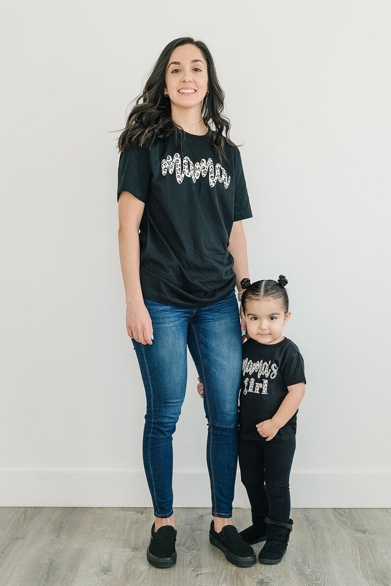 Pode incluir: Uma m&atilde;e e sua filha vestindo camisetas pretas combinando com um design de estampa de leopardo. A camiseta da m&atilde;e diz "Mama" e a camiseta da filha diz "Mama's Girl". Ambas est&atilde;o usando jeans azul e sapatos pretos.