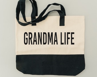 May include: A white canvas tote bag with black handles and a black bottom. The bag has the text "Grandma Life" printed in black on the front.