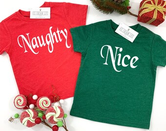 May include: Two short-sleeved t-shirts, one red and one green, with the words "Naughty" and "Nice" in white script. The shirts are displayed with holiday decorations, including a sprig of pine and a glittery gift box.