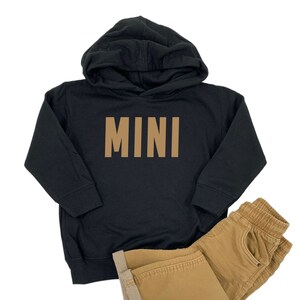 May include: Black hooded sweatshirt with the word "MINI" printed in brown on the front. The sweatshirt is paired with brown pants.