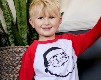 May include: A red and white raglan-style long-sleeve shirt featuring a black and white Santa Claus graphic. The shirt is worn by a young child with blonde hair, smiling at the camera. A large green plant is in the background.