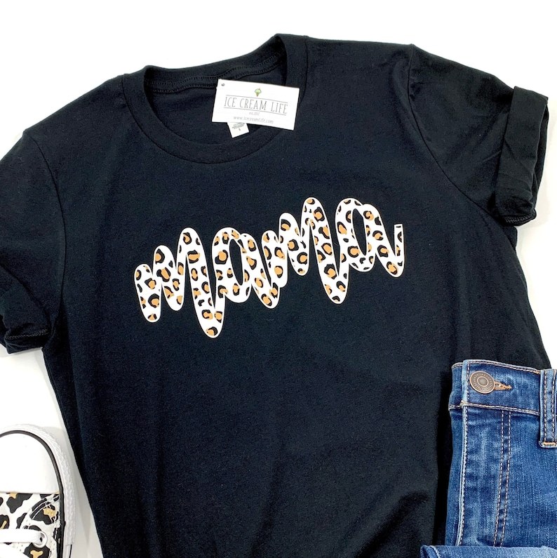 May include: A black t-shirt with a white 'mama' graphic design. The graphic design is in a leopard print pattern with brown and white spots.