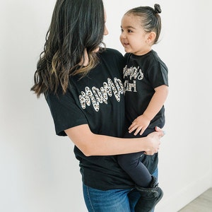 Pode incluir: Uma mulher e uma menina pequena vestindo camisetas pretas combinando. A camiseta da mulher tem um design de estampa de leopardo e o texto "Mama" em branco. A camiseta da menina tem o texto "Mama's Girl" em branco.