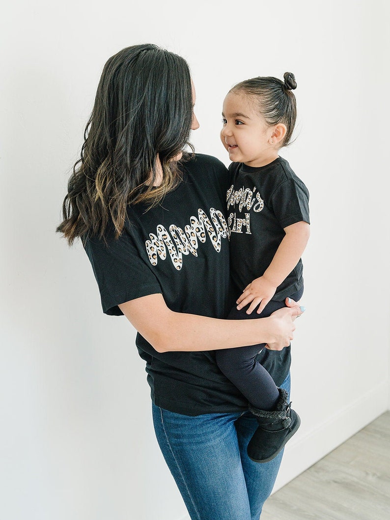 Pode incluir: Uma mulher e uma menina pequena vestindo camisetas pretas combinando. A camiseta da mulher tem um design de estampa de leopardo e o texto "Mama" em branco. A camiseta da menina tem o texto "Mama's Girl" em branco.