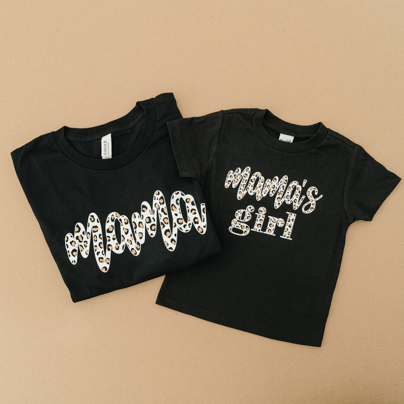 Pode incluir: Duas camisetas pretas com um design de estampa de leopardo. A camiseta maior diz "mama" e a camiseta menor diz "mama's girl".