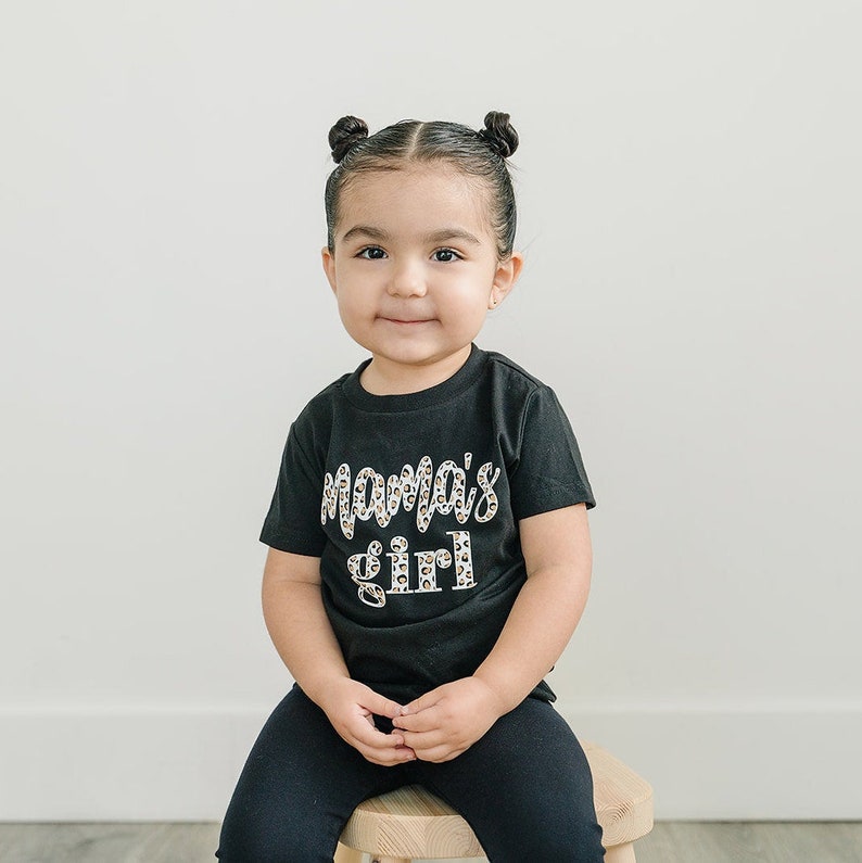 Pode incluir: Uma crian&ccedil;a pequena vestindo uma camiseta preta com um gr&aacute;fico branco "mama's girl" em um design de leopardo. A crian&ccedil;a est&aacute; sentada em um banquinho de madeira e tem o cabelo puxado para tr&aacute;s em duas tran&ccedil;as.