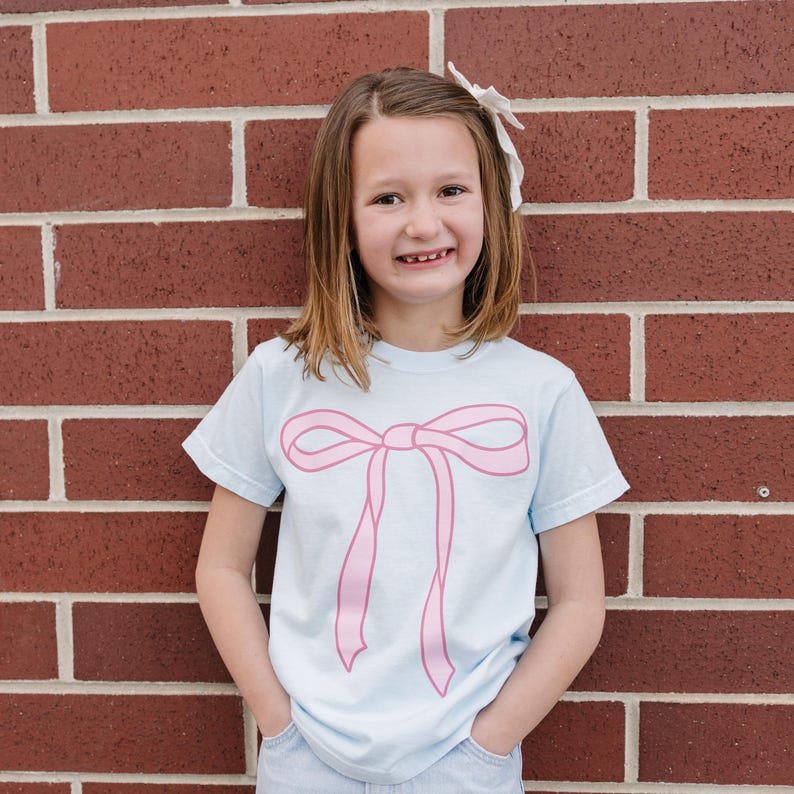 May include: A light blue t-shirt with a pink bow graphic. The shirt is made of a soft, comfortable material.