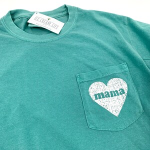 May include: Teal blue t-shirt with a pocket on the front. The pocket has a white heart with the word "mama" inside.