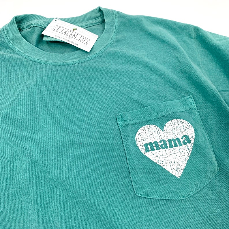 May include: Teal blue t-shirt with a pocket on the front. The pocket has a white heart with the word "mama" inside.