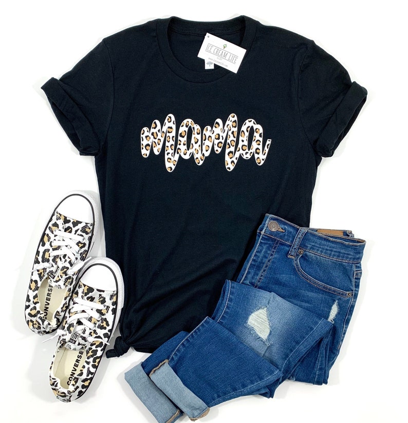 May include: Black t-shirt with a white "mama" graphic in a leopard print design. The shirt is paired with blue jeans and leopard print sneakers.
