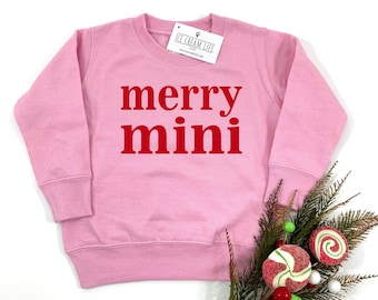 May include: A light pink sweatshirt with the words "merry mini" in red block letters. The sweatshirt has long sleeves and a crew neck. A tag is attached to the neckline. Decorative holiday elements are in the bottom right corner.