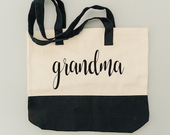 May include: A white canvas tote bag with black handles and a black bottom. The bag has the word "grandma" written in black cursive on the front.