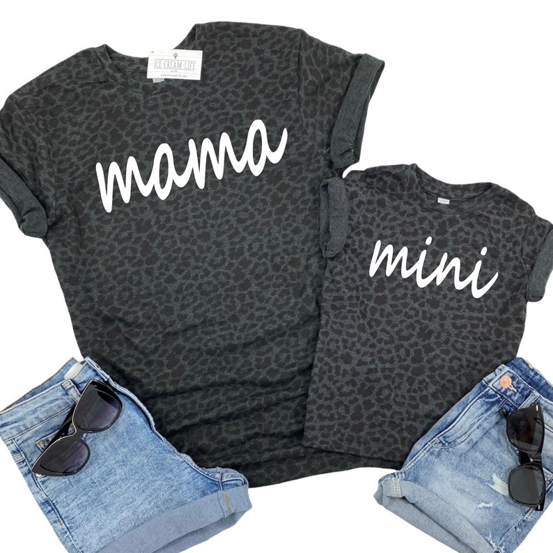 May include: A matching set of two t-shirts with a black leopard print. The larger shirt says "mama" in white script and the smaller shirt says "mini" in white script.
