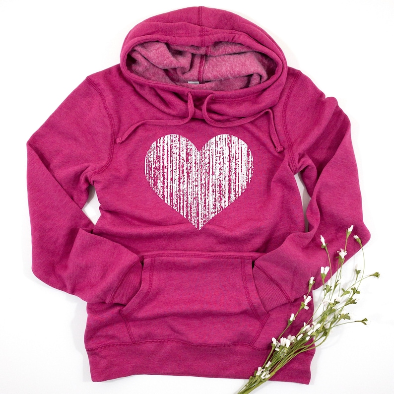 May include: A magenta hoodie with a white, distressed heart graphic. The hoodie features a drawstring hood, a front pocket, and long sleeves. A sprig of white flowers is in the lower right corner. The sweatshirt is made of a soft material.