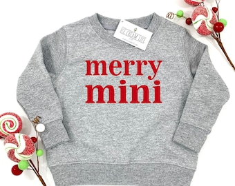 May include: A gray sweatshirt with the words "merry mini" in red block letters. The sweatshirt has a crew neck and long sleeves. A tag that says "ICE CREAM LIFE" is attached to the collar. Decorative candy canes are in the background.