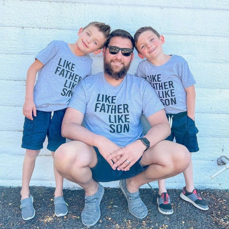 May include: A father and his two sons are wearing matching gray t-shirts with the text "Like Father Like Son". The father is wearing a black watch and a ring on his finger. The boys are wearing black shorts and gray sneakers. The father is wearing black shorts and gray sneakers.