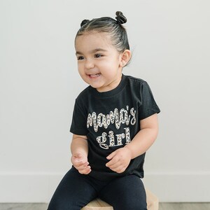 Pode incluir: Uma crian&ccedil;a pequena vestindo uma camiseta preta com as palavras "Mama's girl" impressas em branco com um padr&atilde;o de leopardo. A crian&ccedil;a est&aacute; sentada em um banquinho de madeira.
