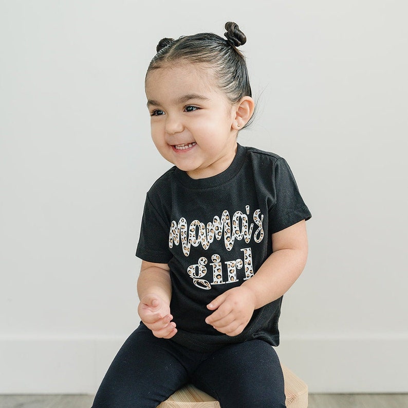 Pode incluir: Uma crian&ccedil;a pequena vestindo uma camiseta preta com as palavras "Mama's girl" impressas em branco com um padr&atilde;o de leopardo. A crian&ccedil;a est&aacute; sentada em um banquinho de madeira.