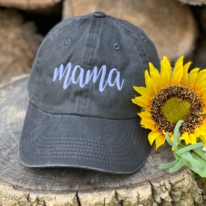 May include: A black baseball cap with a white embroidered word "mama" on the front. The cap is sitting on a wooden surface with a yellow sunflower.