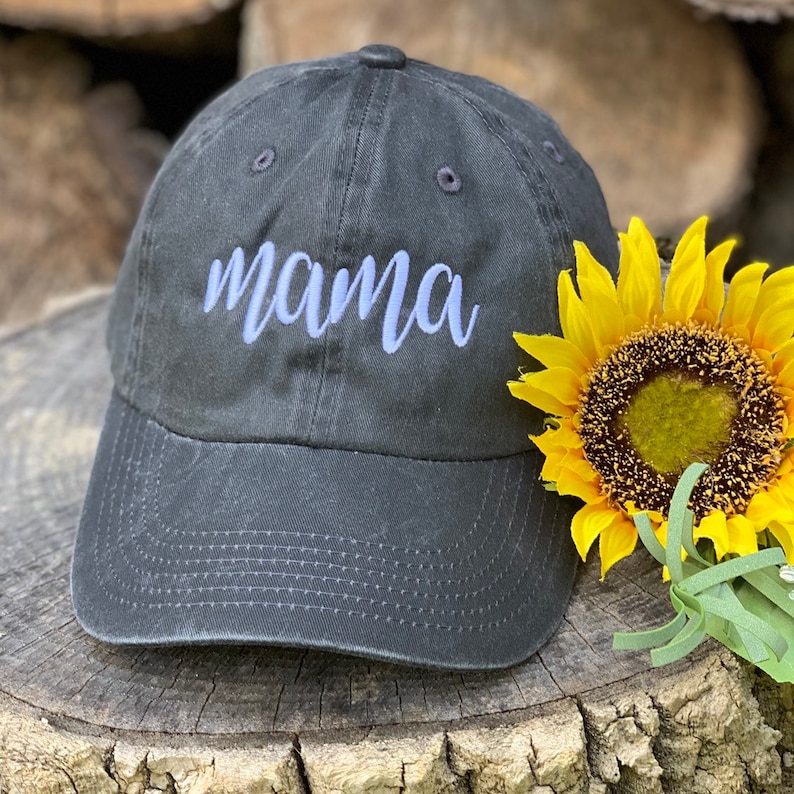 May include: A black baseball cap with a white embroidered word "mama" on the front. The cap is sitting on a wooden surface with a yellow sunflower.