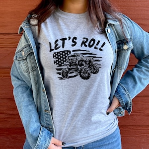 Puede incluir: Una camiseta gris con el texto "Let's Roll" impreso en ella. El texto está en negro y está rodeado por un diseño de bandera estadounidense en blanco y negro. Una silueta negra de un vehículo todoterreno lado a lado está en el centro de la bandera.