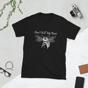 May include: Black t-shirt with a white graphic of a bee and the text "Don't Kill My Buzz". The design features a detailed illustration of a bee with its wings spread. The shirt is made of a soft material.