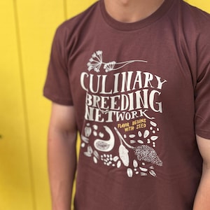 May include: A person wearing a brown t-shirt with the text "Culinary Breeding Network" and the phrase "Flavor Begins With Seed" printed on the front. The shirt features a white graphic design of various seeds and plants.