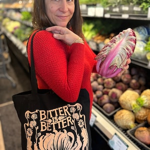 May include: A black tote bag with the text "Bitter is Better" printed in gold. The bag features a stylized illustration of a leafy vegetable with a floral border. A woman is holding the bag and a large red radicchio.