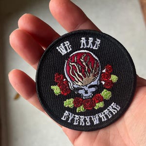 May include: A black embroidered patch with a skull and flames in the center. The skull is surrounded by red roses and the text "WE ARE EVERYWHERE" is written around the skull.