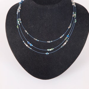 May include: A three-strand necklace with blue, green, and silver beads. The necklace is on a black velvet display stand.