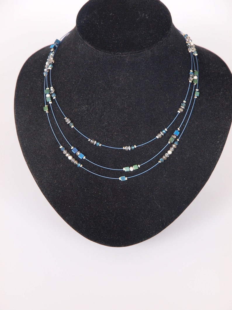 May include: A three-strand necklace with blue, green, and silver beads. The necklace is on a black velvet display stand.