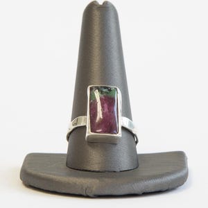 May include: A silver ring with a rectangular, multi-colored stone, primarily red and green, is displayed on a dark gray ring holder. The ring's band is a simple, polished silver. The stone's colors create a striking contrast against the silver.
