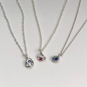 Puede incluir: Tres collares de plata con diferentes diseños de colgantes. Uno tiene una gema azul, otro una gema roja y el tercero una gema azul engastada en un colgante circular. Todos los collares tienen delicadas cadenas de plata.