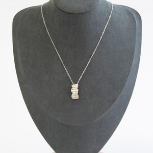 May include: A silver necklace with a pendant featuring a stacked, textured design. The necklace is displayed on a gray mannequin, showcasing the delicate chain and the pendant's unique, layered appearance.