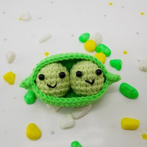 Pu&ograve; includere: Un baccello di piselli all'uncinetto fatto a mano con due piselli sorridenti. Il baccello &egrave; verde brillante e i piselli sono verde chiaro con occhi neri e bocche sorridenti. Lo sfondo &egrave; bianco con elementi decorativi colorati sparsi.