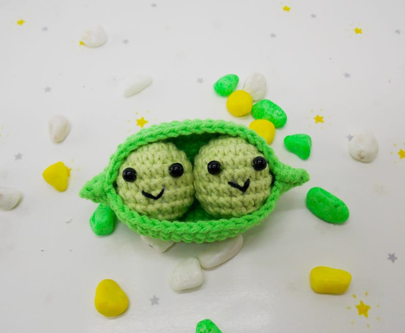 Pu&ograve; includere: Un baccello di piselli all'uncinetto fatto a mano con due piselli sorridenti. Il baccello &egrave; verde brillante e i piselli sono verde chiaro con occhi neri e bocche sorridenti. Lo sfondo &egrave; bianco con elementi decorativi colorati sparsi.