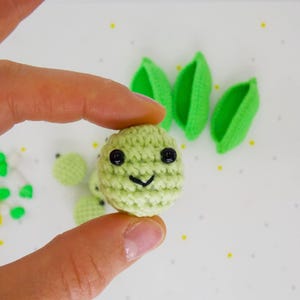 Pu&ograve; includere: Un viso sorridente verde chiaro all'uncinetto con occhi neri e bocca curva &egrave; tenuto tra le dita. Diverse altre facce all'uncinetto e forme di foglie verdi sono sparse nelle vicinanze. Lo sfondo &egrave; bianco con piccoli punti gialli e grigi.