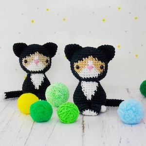 May include: Two black and white crocheted cat toys with pink noses and orange and white markings on their faces. The cats are sitting on a white wooden surface with green, yellow, and blue yarn balls.