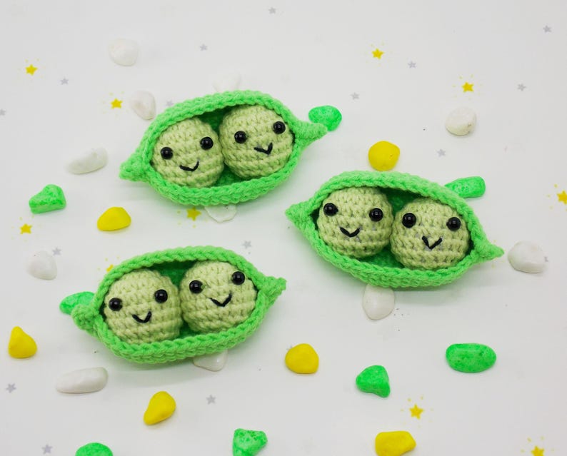 Pu&ograve; includere: Tre baccelli di piselli all'uncinetto, ciascuno con due piselli sorridenti. I piselli sono verde chiaro con occhi neri e sorrisi cuciti. I baccelli sono verde brillante. Piccole forme decorative gialle, verdi e bianche sono sparse.